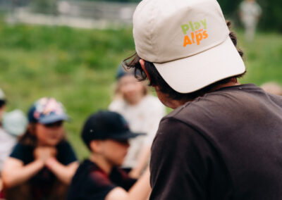 Eine Person, von hinten gesehen, mit einer „Play in the Alps“-Kappe, die den Kindern Anweisungen gibt.
